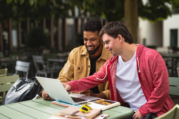 A young man with Down syndrome sitting outdoors with his mentoring friend using a laptop together.